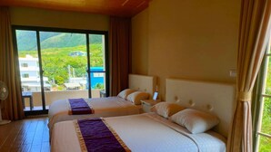 A hotel room with two beds, a balcony with a view of greenery and buildings, and a fan on the floor. - Two Fish (Lanyu)