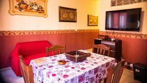 Un coin repas avec une table recouverte d’une nappe à motifs, un canapé rouge et un téléviseur fixé au mur.