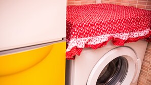 Une machine à laver recouverte d’une housse à motifs rouges et blancs, avec un meuble jaune et un mur carrelé.
