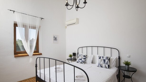 A bedroom with a bed, a window with sheer curtains, a bedside table with a lamp, and a small plant.