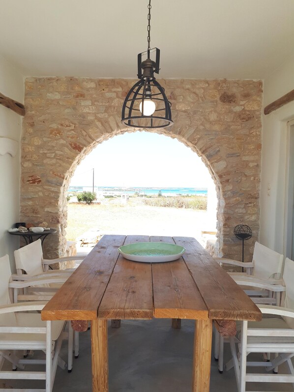 A wooden table with chairs in a stone-walled room with an arched doorway leading to a view of the sea.
