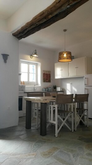 A kitchen with a stone floor, wooden table, and chairs, a hanging light fixture, and a window.