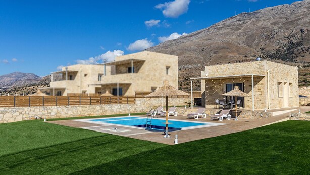Zona de piscina con edificio de piedra, césped y montañas al fondo.