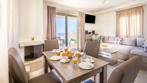 Un comedor con una mesa preparada para el desayuno, incluyendo platos, vasos y un bol de fruta. Hay un sofá y una televisión al fondo.