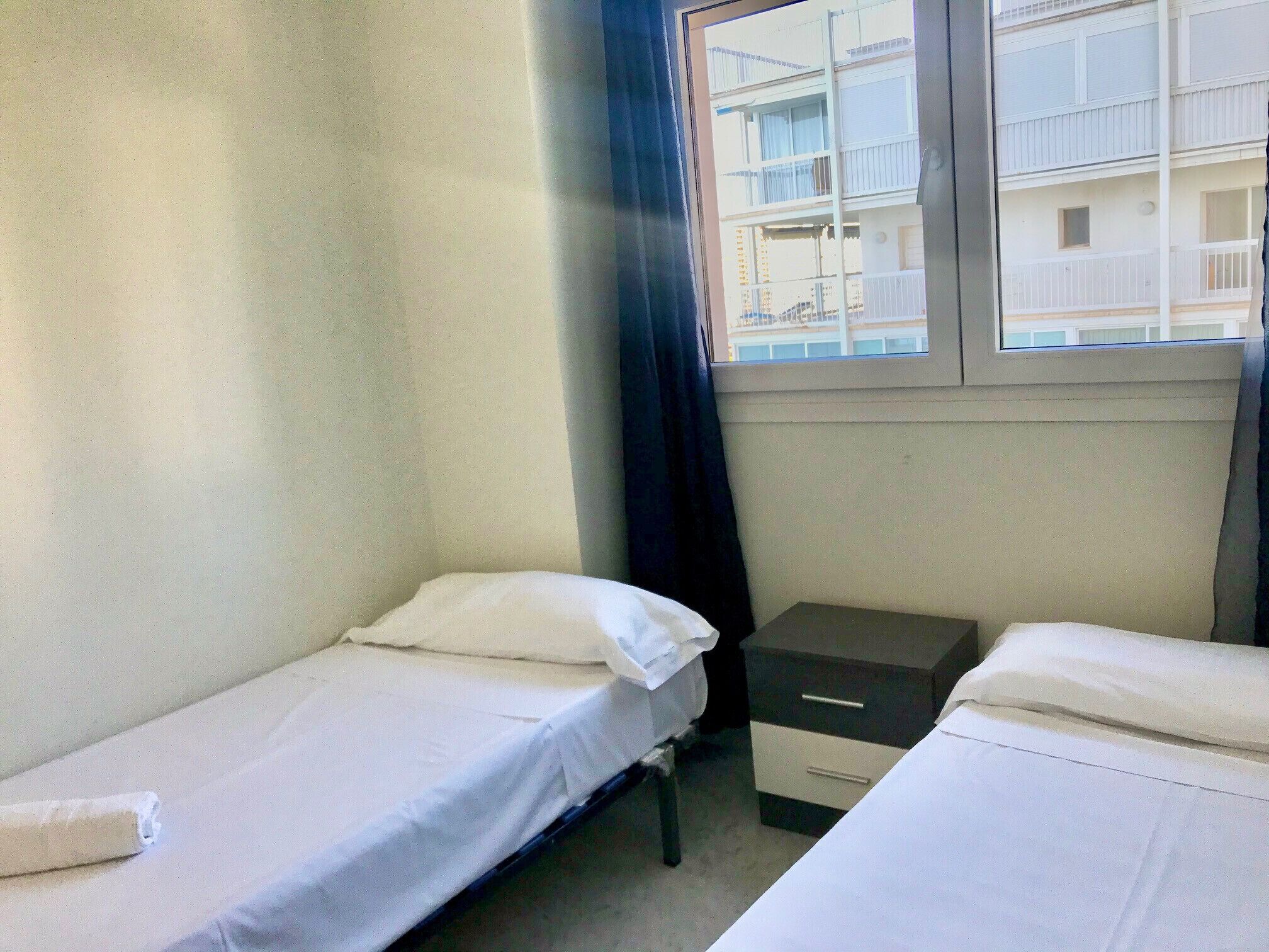 A small, single-room hotel room with two single beds, a bedside table, and a window with a view of a building.