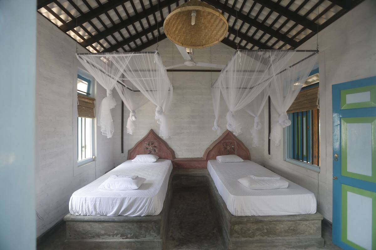 A room with two beds, each with white bedding and a canopy. The room has a high ceiling with a woven ceiling fan.