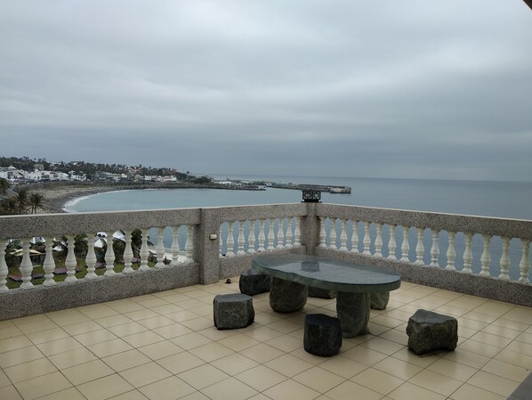 Une terrasse avec une table et des sièges, offrant une vue sur la côte, avec une promenade et des bâtiments visibles au loin.