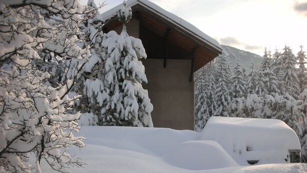 A snow-covered building with a sloped roof, surrounded by snow-laden trees and mountains in the background.