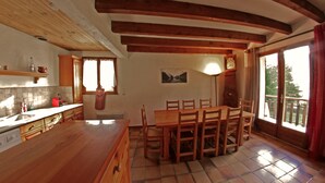 A kitchen with wooden beams, a dining table with chairs, a window with curtains, and a clock on the wall.