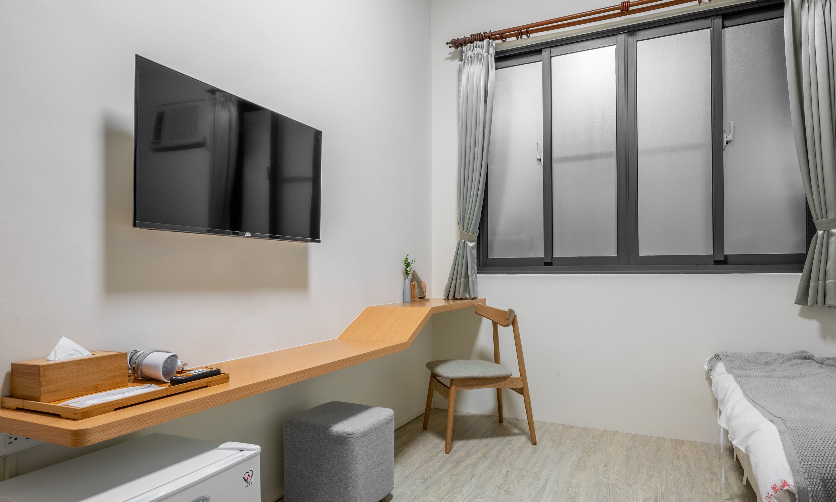 Une chambre moderne avec un téléviseur fixé au mur, un bureau en bois avec une chaise, un lit recouvert d’une couverture grise et une fenêtre avec des rideaux.
