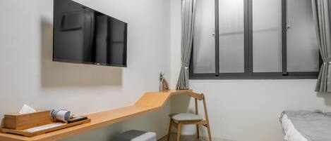 Une chambre moderne avec un téléviseur fixé au mur, un bureau en bois avec une chaise, un lit recouvert d’une couverture grise et une fenêtre avec des rideaux.