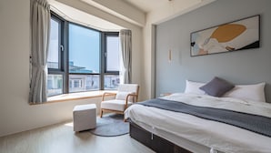 Een moderne slaapkamer met een groot bed, een stoel, een klein voetenbankje en uitzicht op de stad door grote ramen.