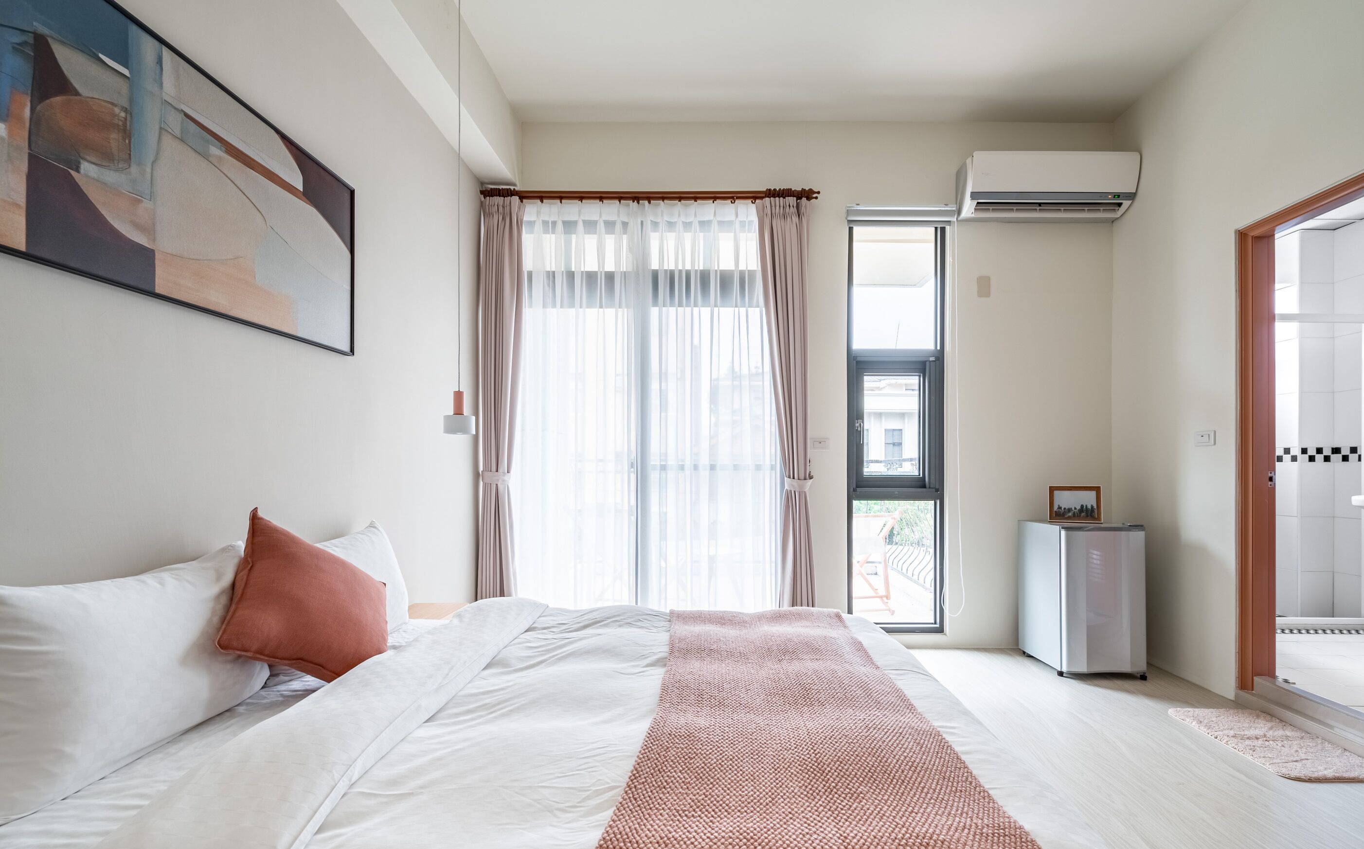 A bedroom with a bed, a window with sheer curtains, an air conditioning unit, and a door leading to another room.