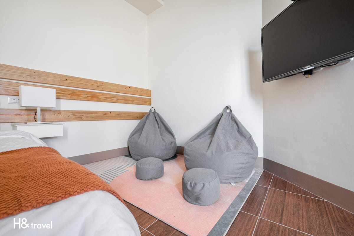 A hotel room with a bed, a TV mounted on the wall, and two bean bags on a pink rug.