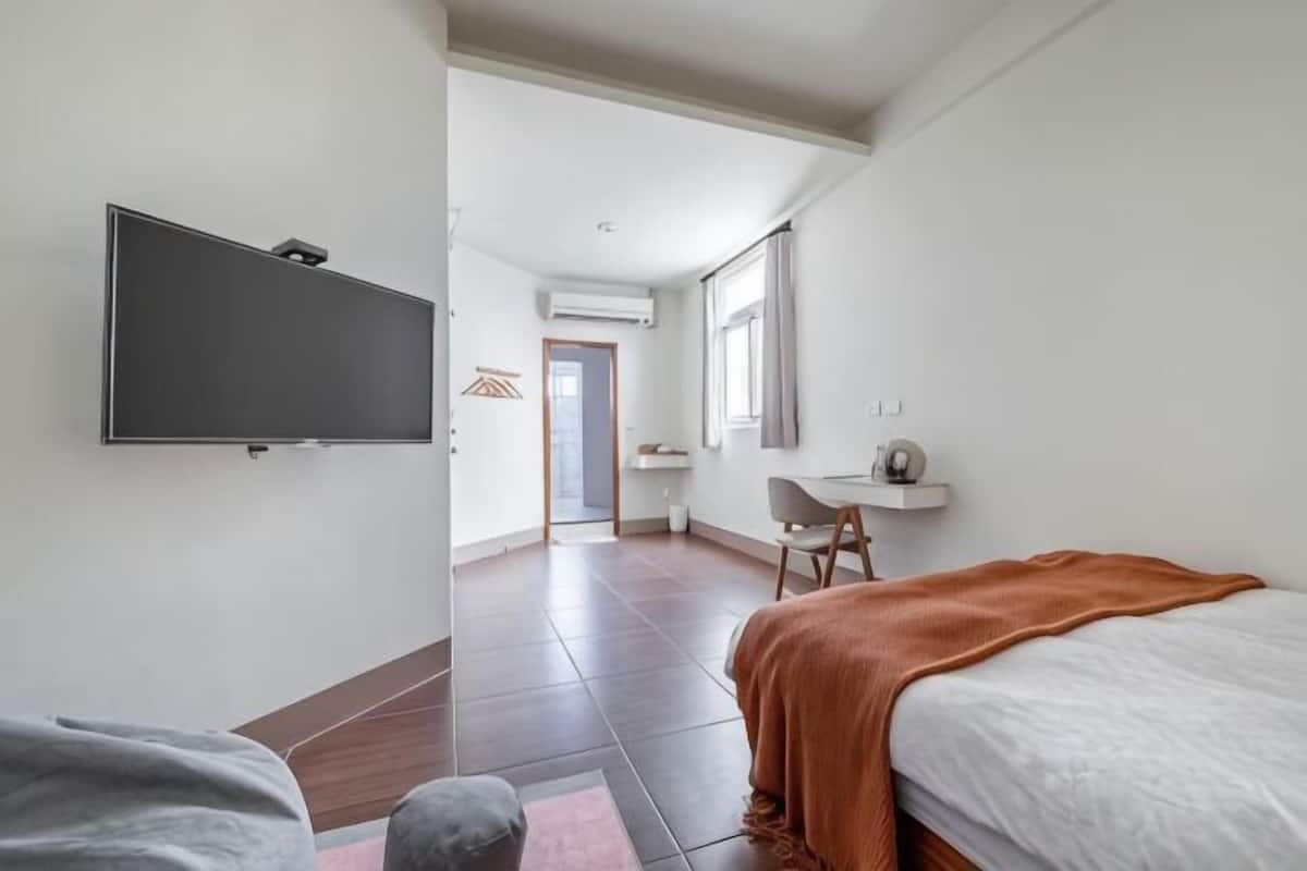 A bedroom with a bed, a desk, a chair, a television, and an air conditioning unit.