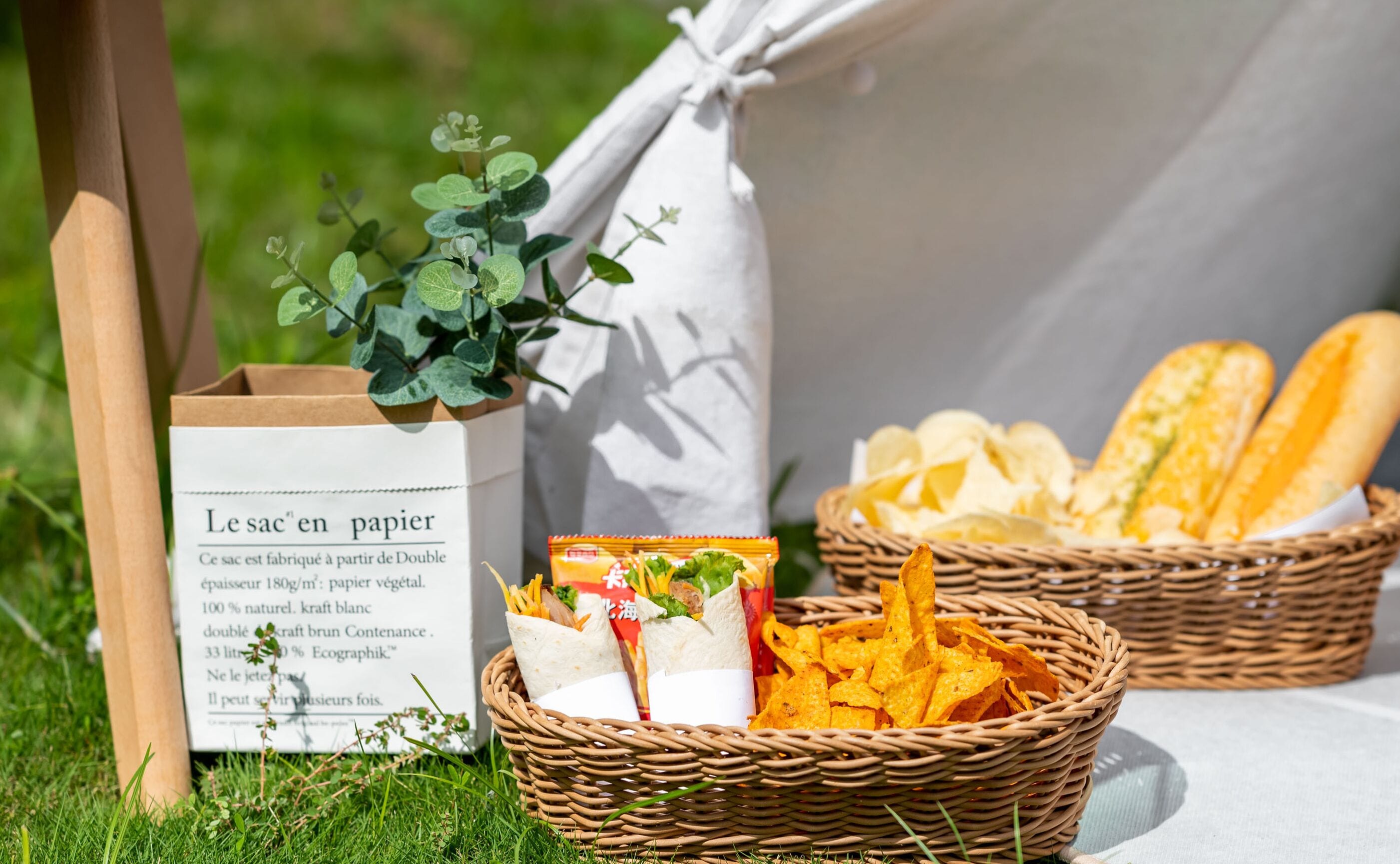 A picnic setup with a paper bag labeled "Le sac en papier," containing food items and a basket with baguettes and chips.