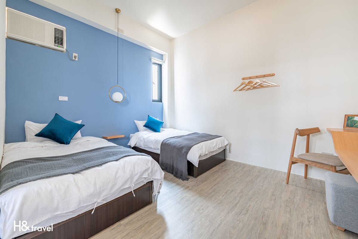 A hotel room with two beds, a wooden chair, a small table, and a wall-mounted air conditioner.