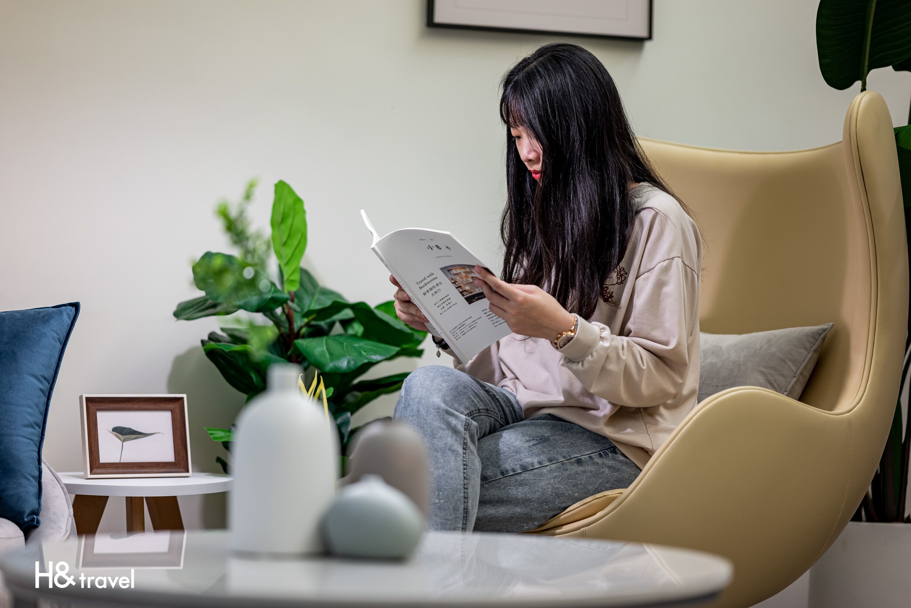 A person sitting on a modern chair reading a book in a well-lit room with a plant and a small table with a framed picture.