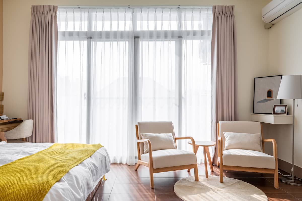 A hotel room with a bed, two chairs, a desk, a lamp, and a large window with sheer curtains.