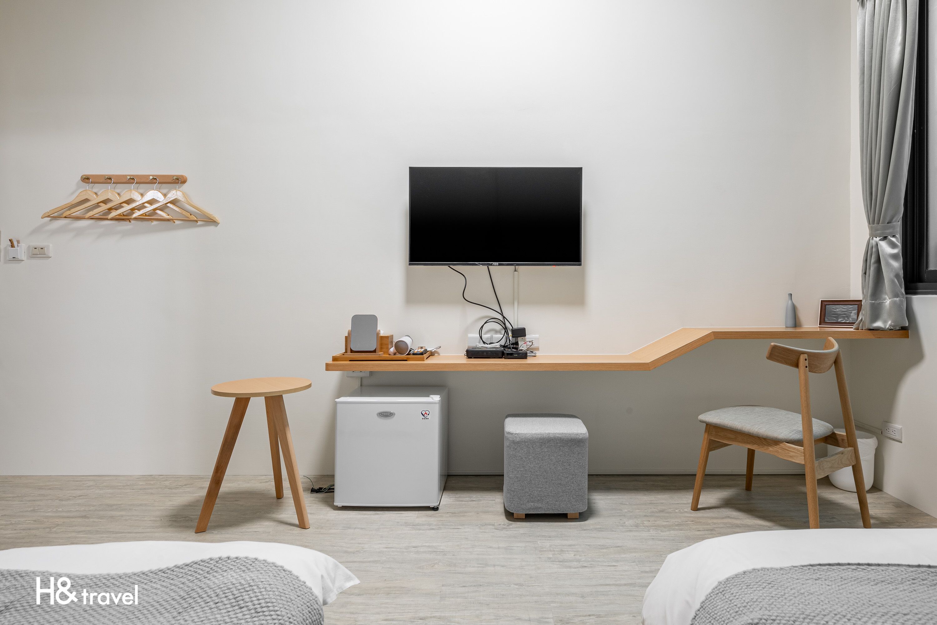 Una habitación de hotel moderna con un escritorio de madera, televisor, mini refrigerador, un otomano gris y una silla de madera.