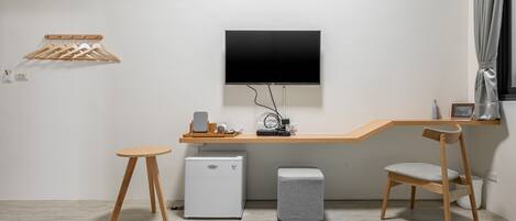 Una habitación de hotel moderna con un escritorio de madera, televisor, mini refrigerador, un otomano gris y una silla de madera.