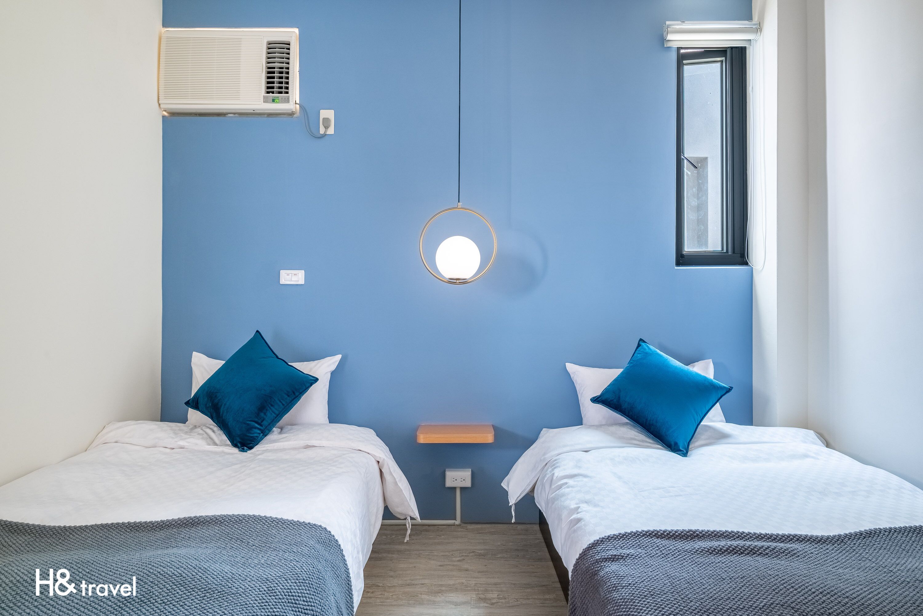 A hotel room with two beds, a round mirror, an air conditioner, a window, and a small table.