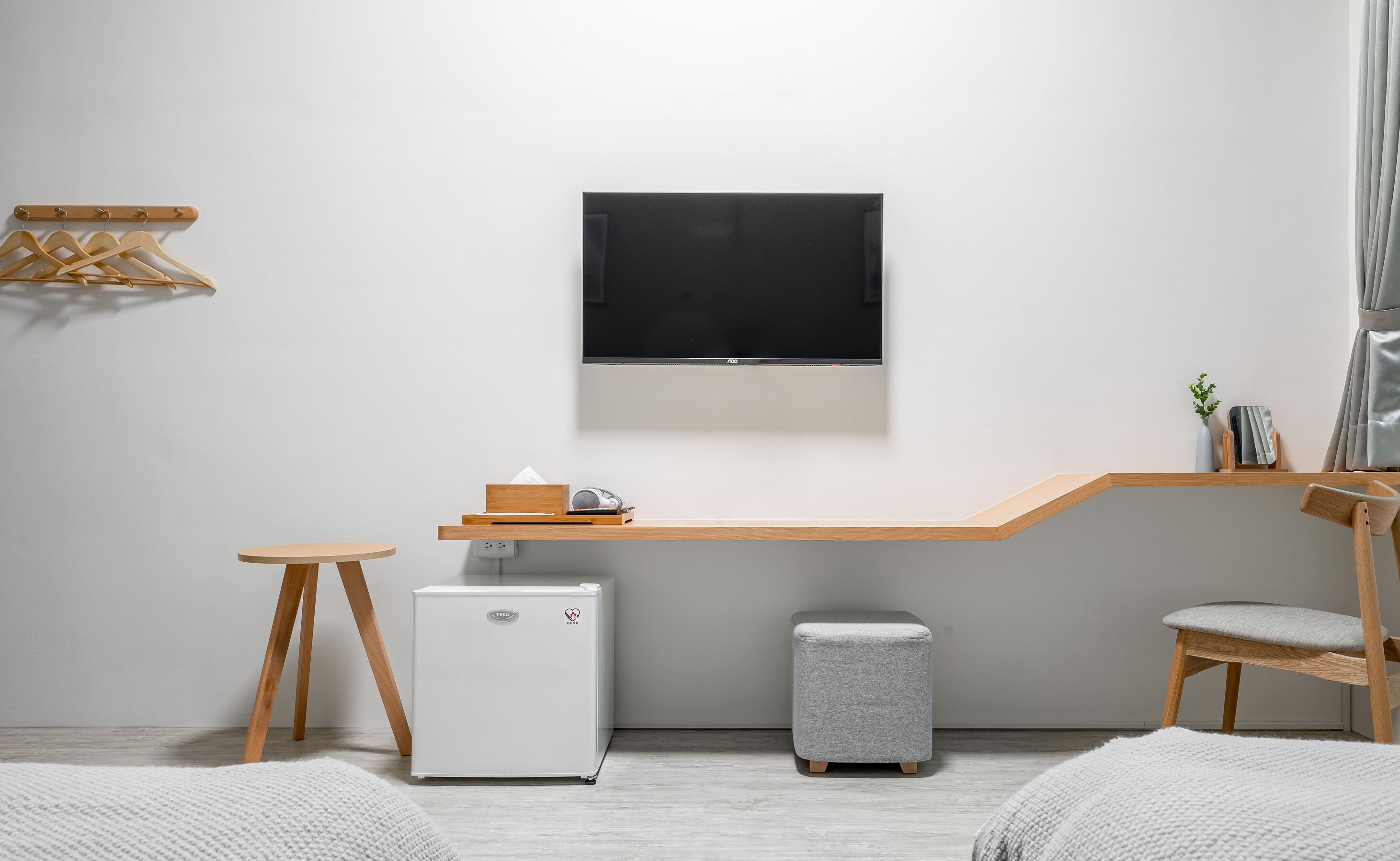 A minimalist room with a flat-screen TV mounted on the wall, a small white refrigerator, a wooden desk with a computer, a grey ottoman, and a wooden chair.