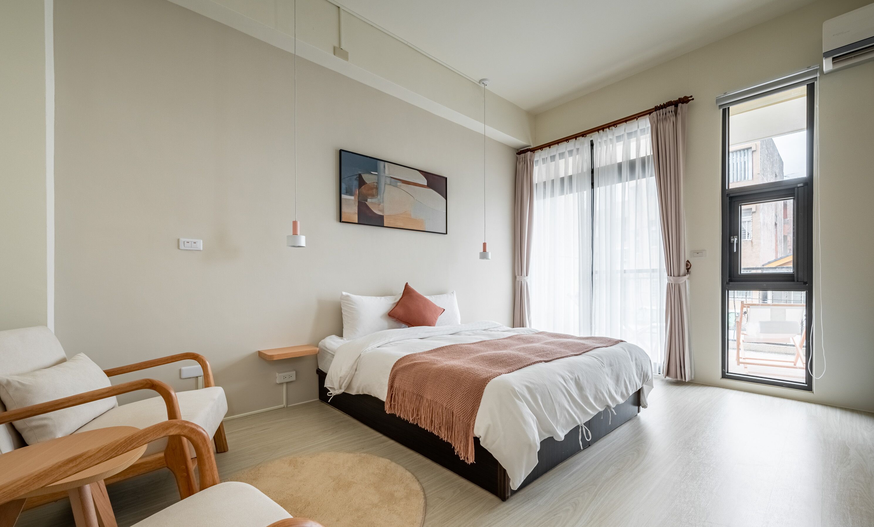 A modern bedroom with a bed, bedside table, and a chair. There is a window with curtains and a view of the outside.