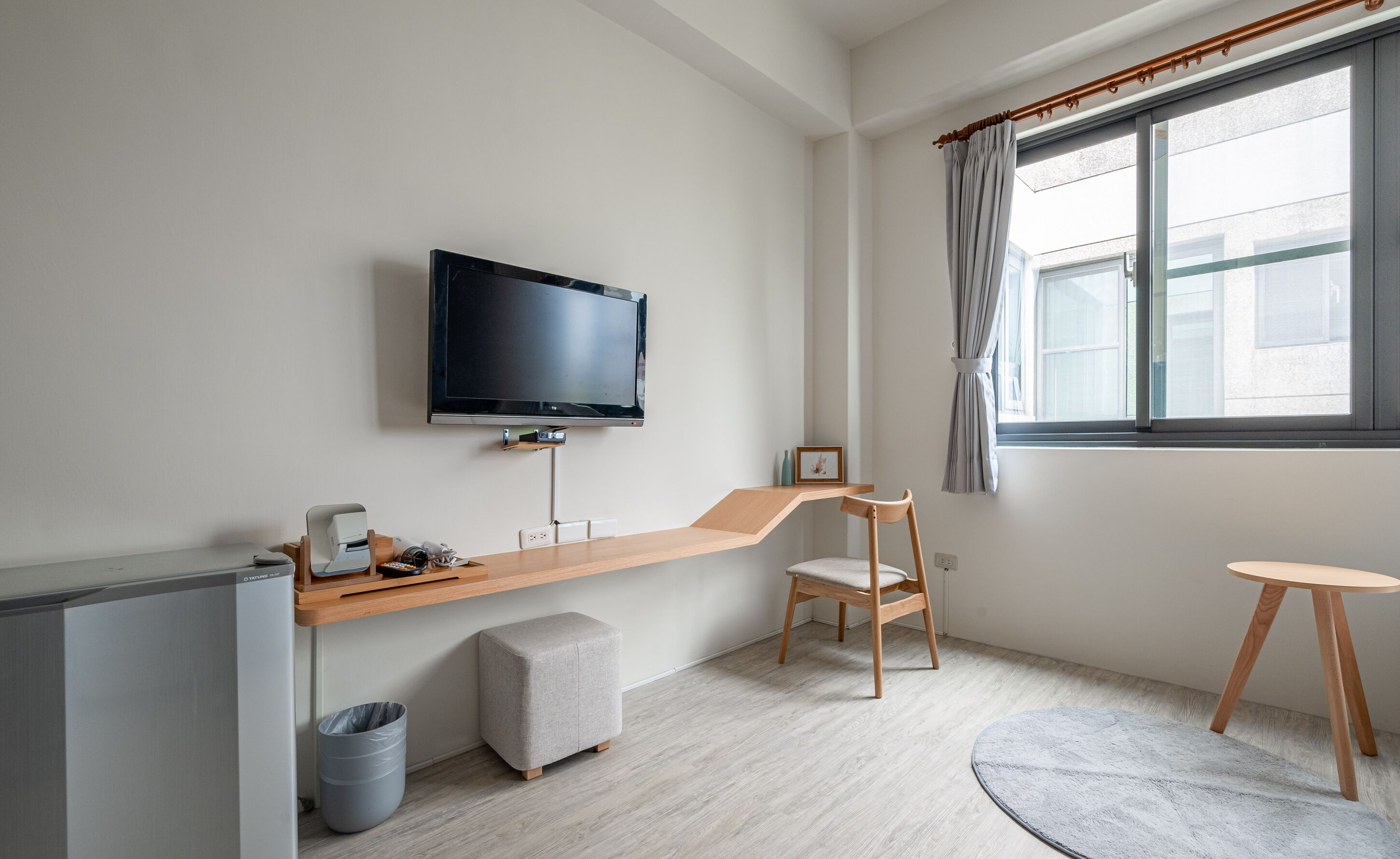 Une chambre d’hôtel moderne équipée d’un téléviseur à écran plat fixé au mur, d’un bureau en bois avec une chaise, d’un petit pouf et d’un tapis rond.
