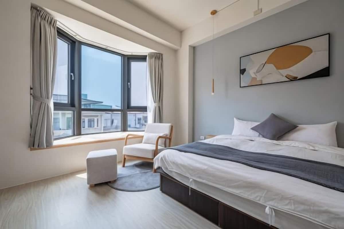 A modern bedroom with a large bed, a chair, a window with curtains, and a city view.