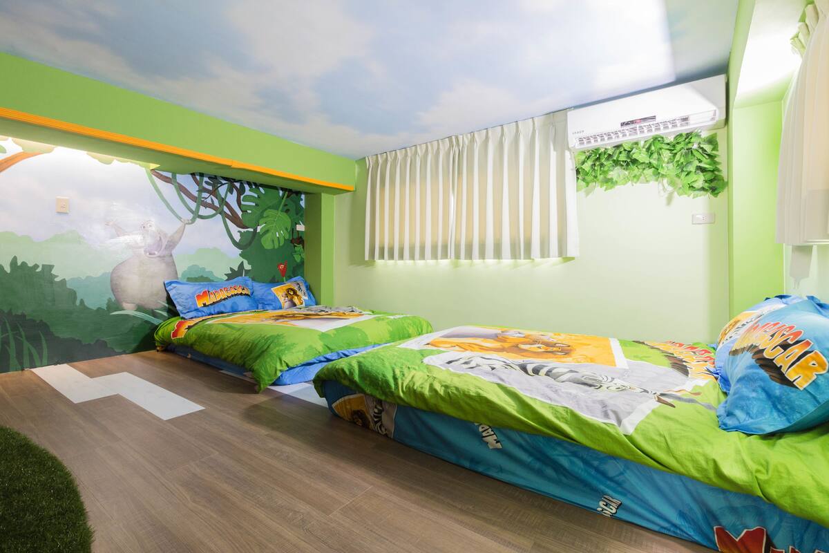 Quarto de criança com duas camas, paredes verdes, um mural de uma cena de selva e uma janela com persianas.