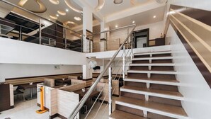 Interior moderno de hotel com uma escadaria central, diversas áreas de refeições e uma saída de emergência visível.