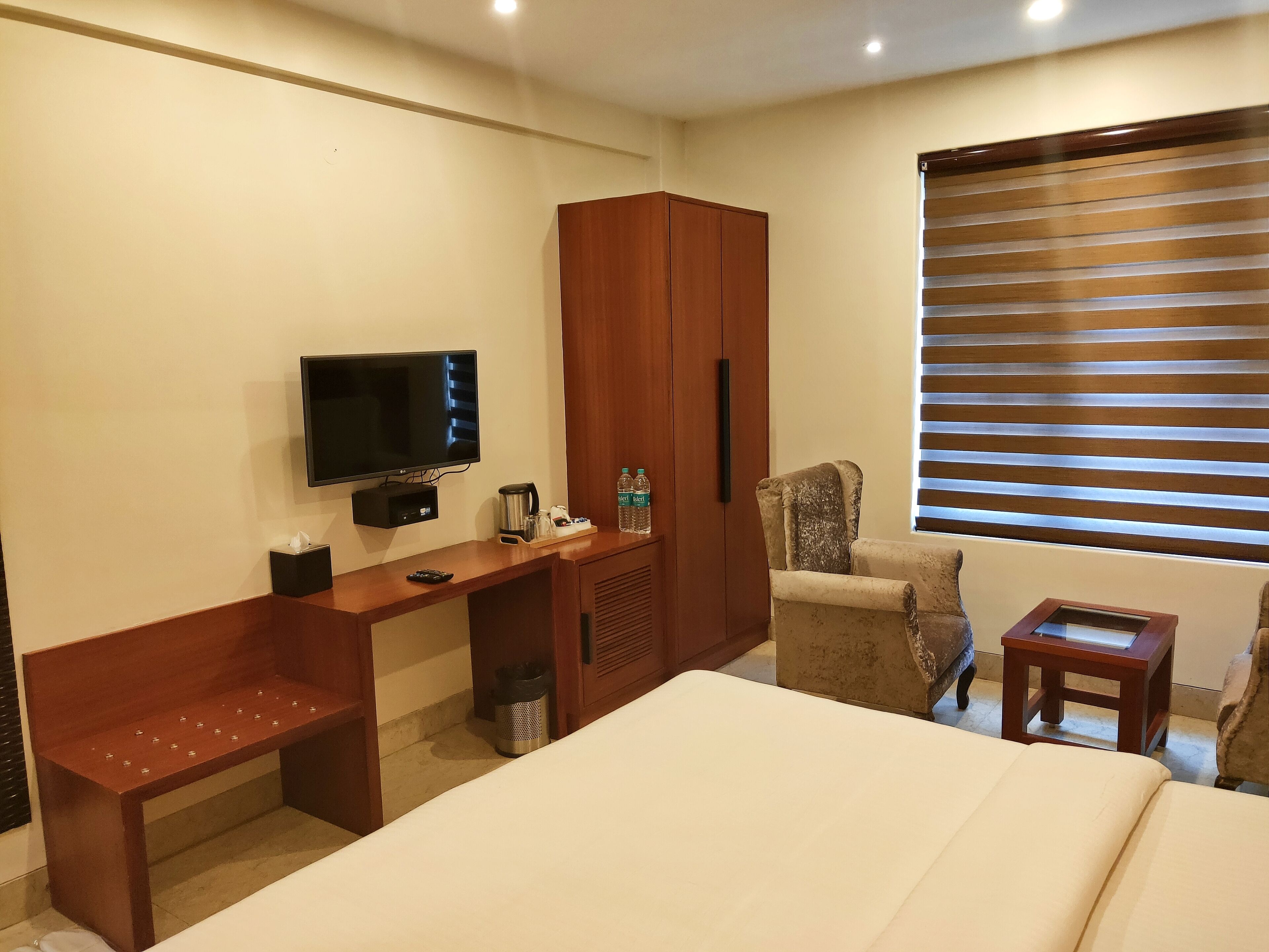 A hotel room with a bed, a wooden desk, a television, a wardrobe, a chair, and a window with blinds.