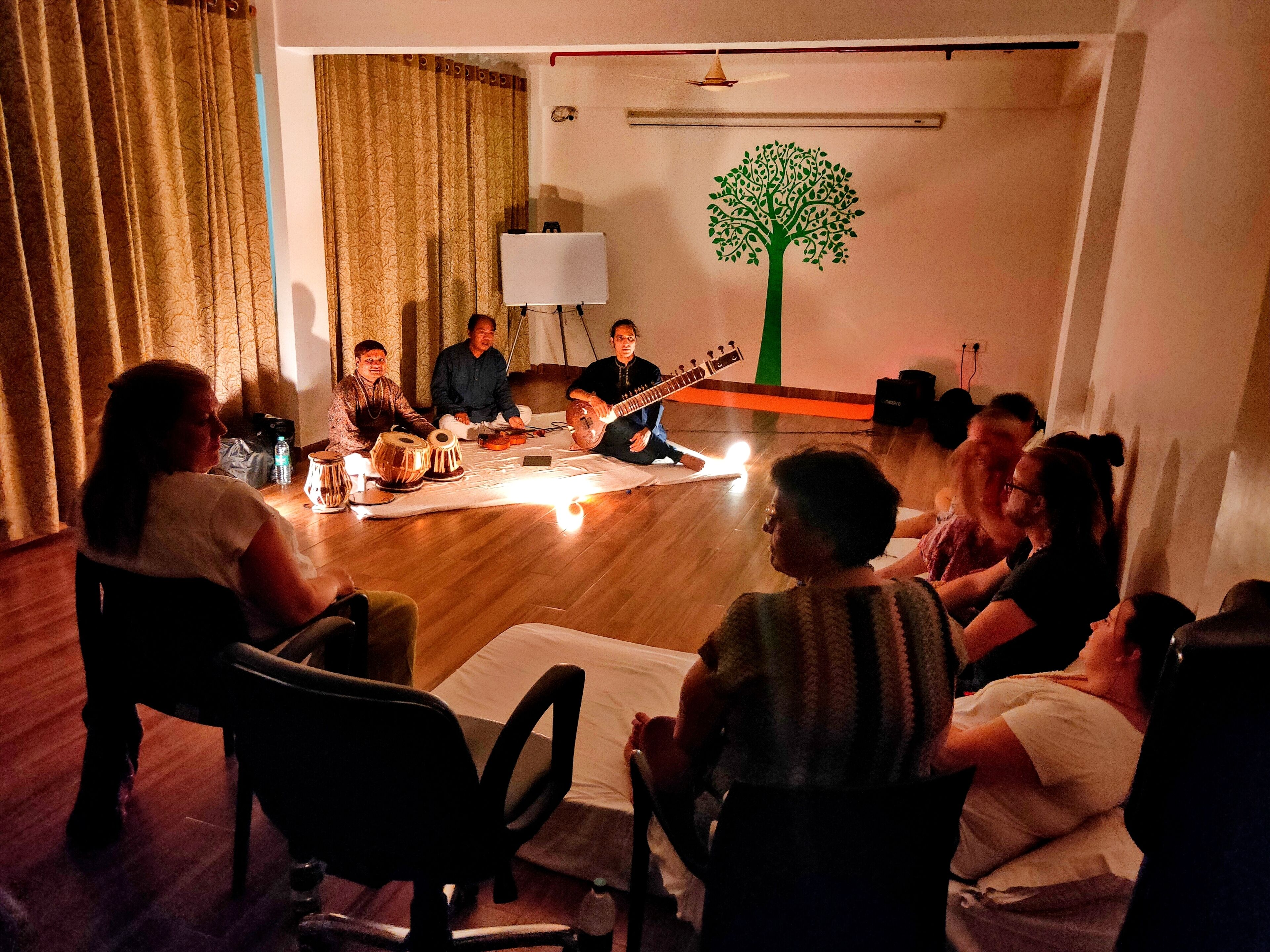 Een groep mensen zitten in een kring op een houten vloer, en kijken naar een muzikant die een snaarinstrument bespeelt.