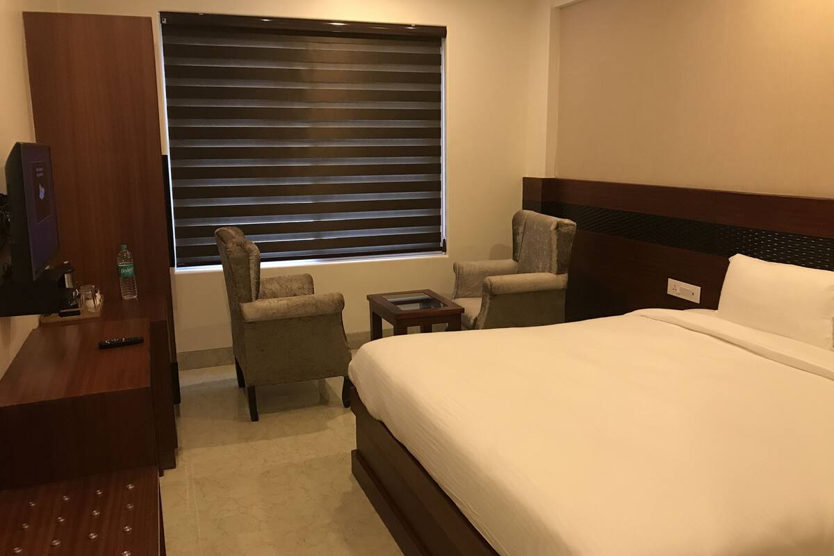 Ein Hotelzimmer mit einem großen Bett, zwei Sesseln, einem kleinen Tisch, einem Fernseher und einem Fenster mit Jalousien.