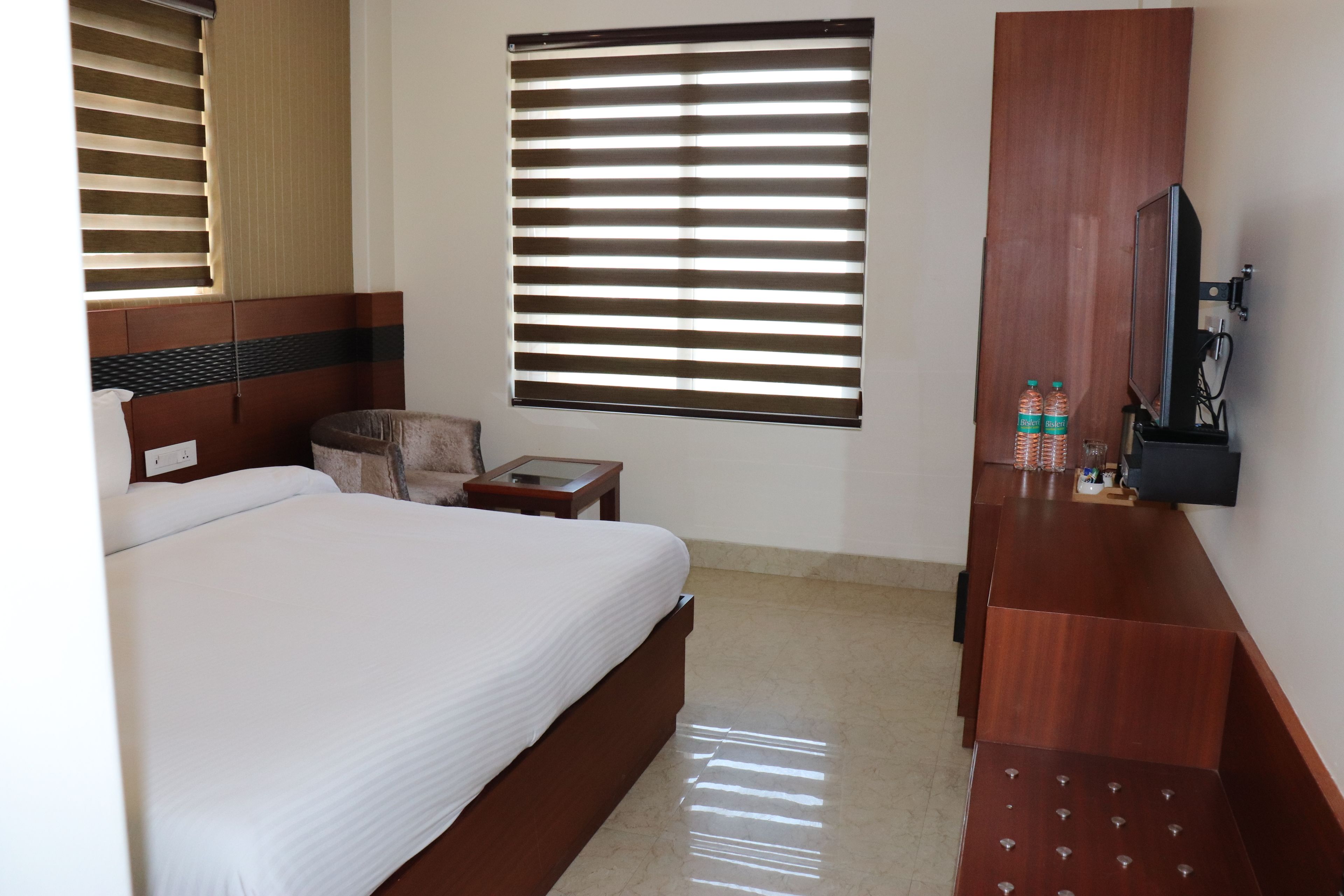 A hotel room with a bed, a wooden desk, a television, and a window with blinds.