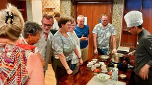 Un grupo de personas reunidas alrededor de una mesa con diferentes alimentos y utensilios de cocina, sugiriendo un evento de degustación o preparación de alimentos en una habitación con suelo de madera y papel tapiz estampado.