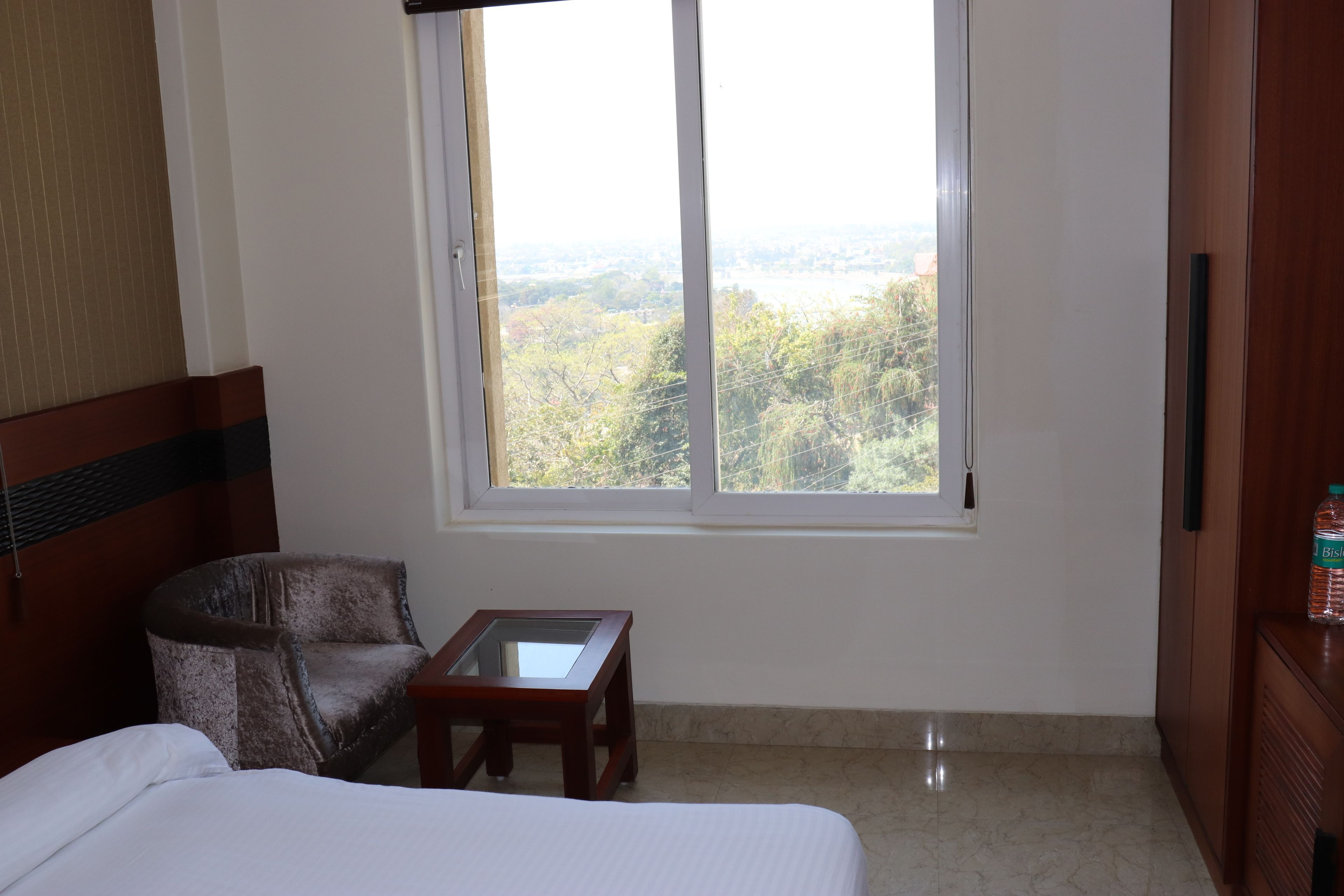 Habitación de hotel con cama, una silla, una mesita y ventana con vista a un área boscosa.