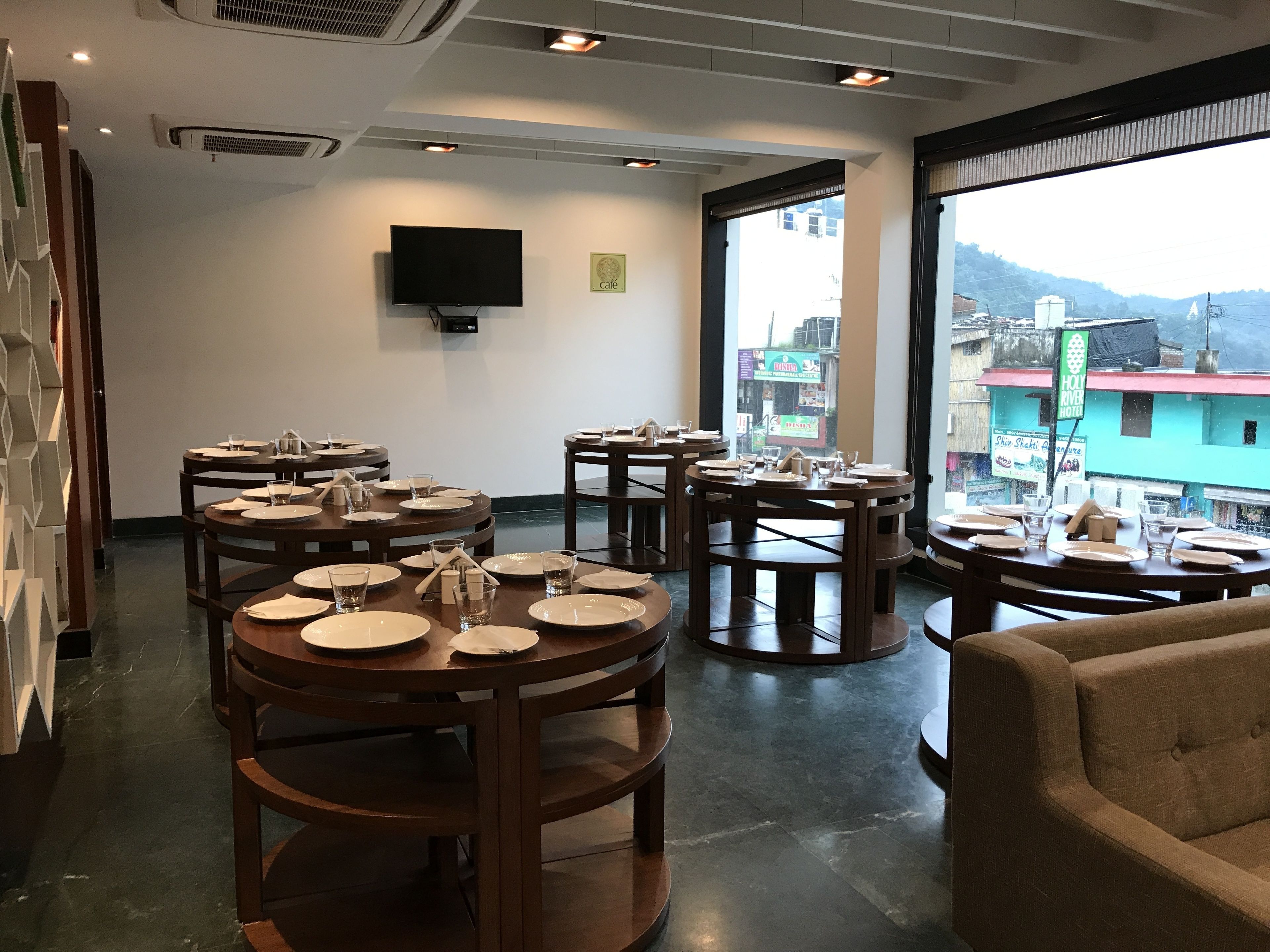 A restaurant with round tables set for dining, a flat-screen TV mounted on the wall, and large windows offering a view of the cityscape.