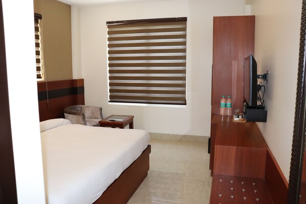 A hotel room with a bed, a small round table, a chair, a wooden dresser, a television, and a window with blinds. - Holy River Hotel (Narendranagar)