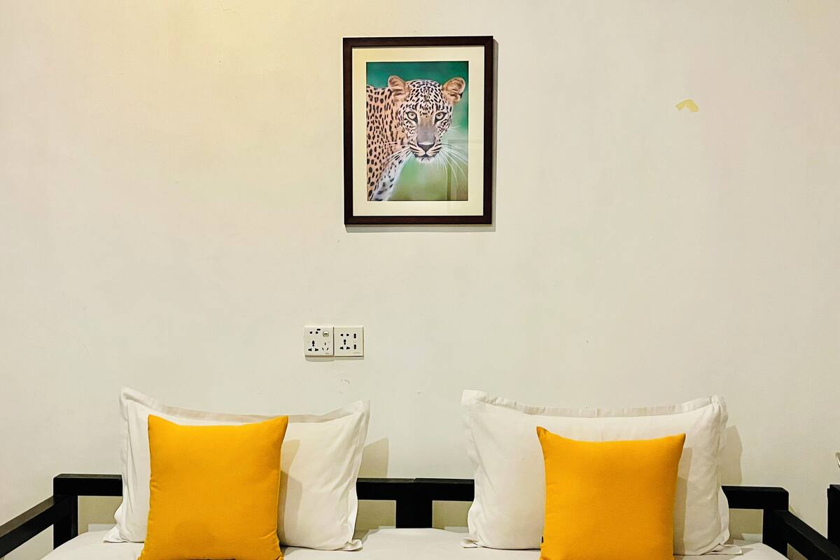 Quarto de hotel com uma cama, roupa de cama branca e dois travesseiros amarelos. Um quadro com a imagem de um leopardo está pendurado na parede acima da cama.