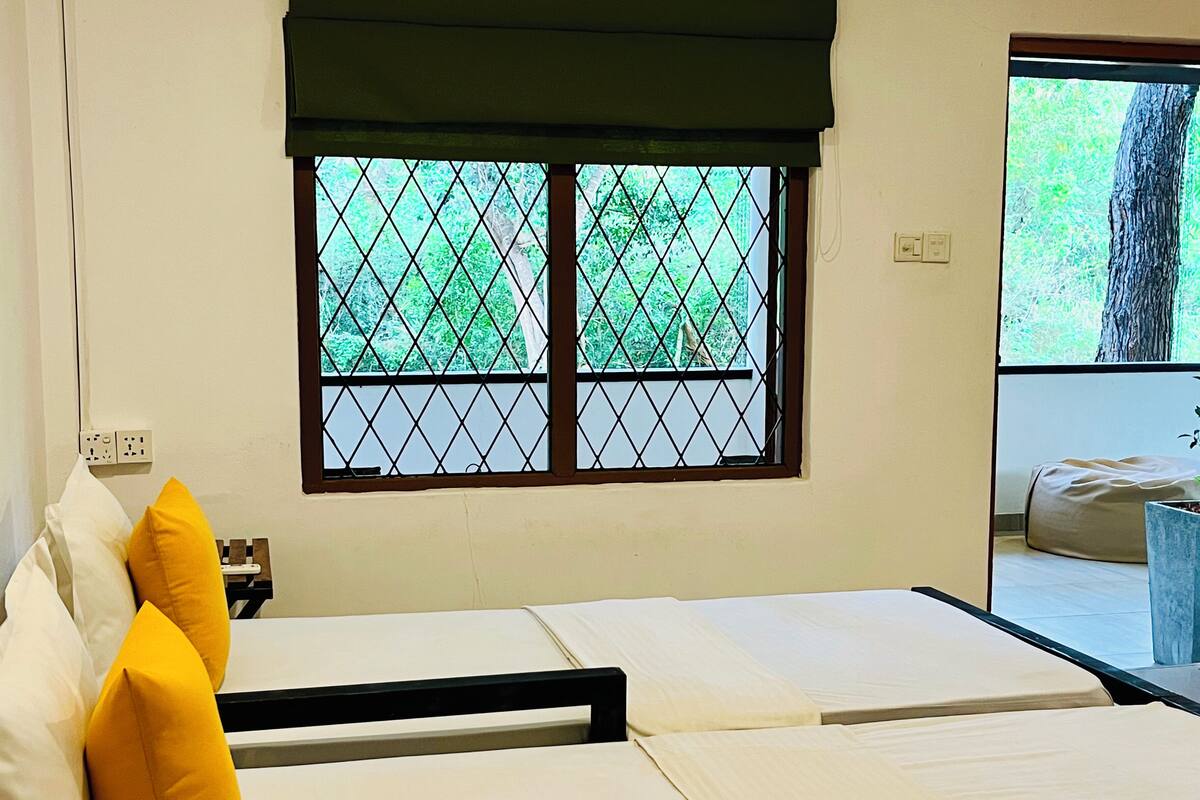 Quarto com cama, almofadas, janela com vista para vegetação e uma tomada na parede.