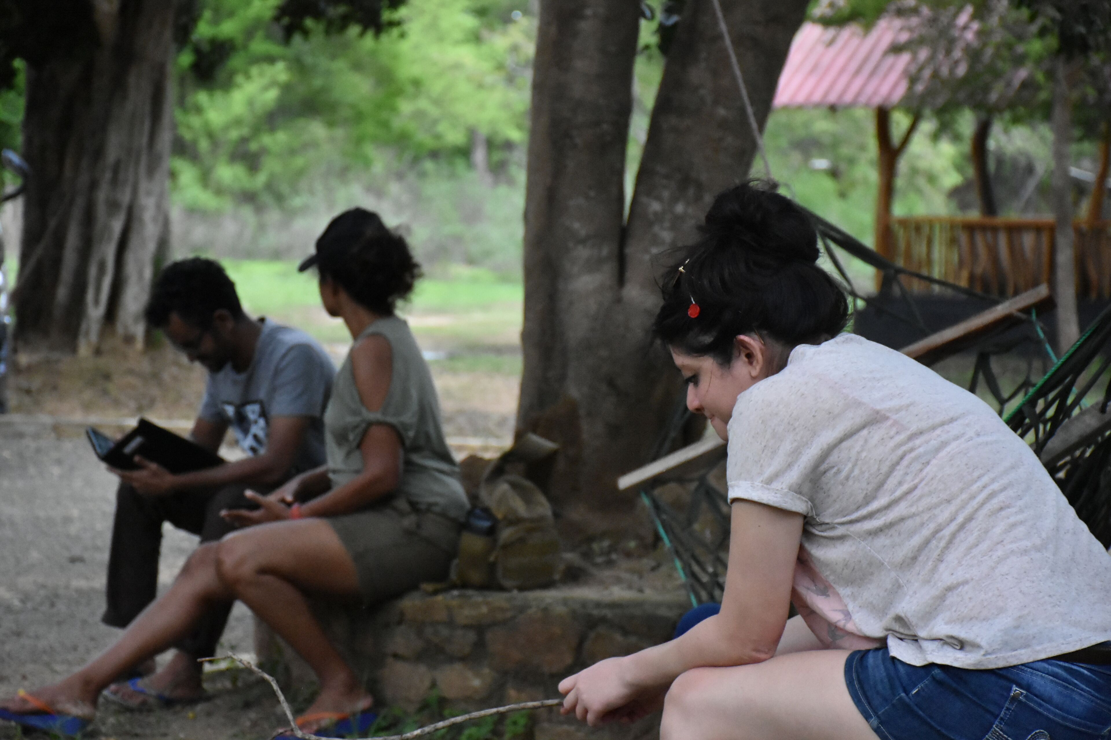 Three individuals sitting on a stone bench in a park-like setting with trees and a wooden structure in the background.