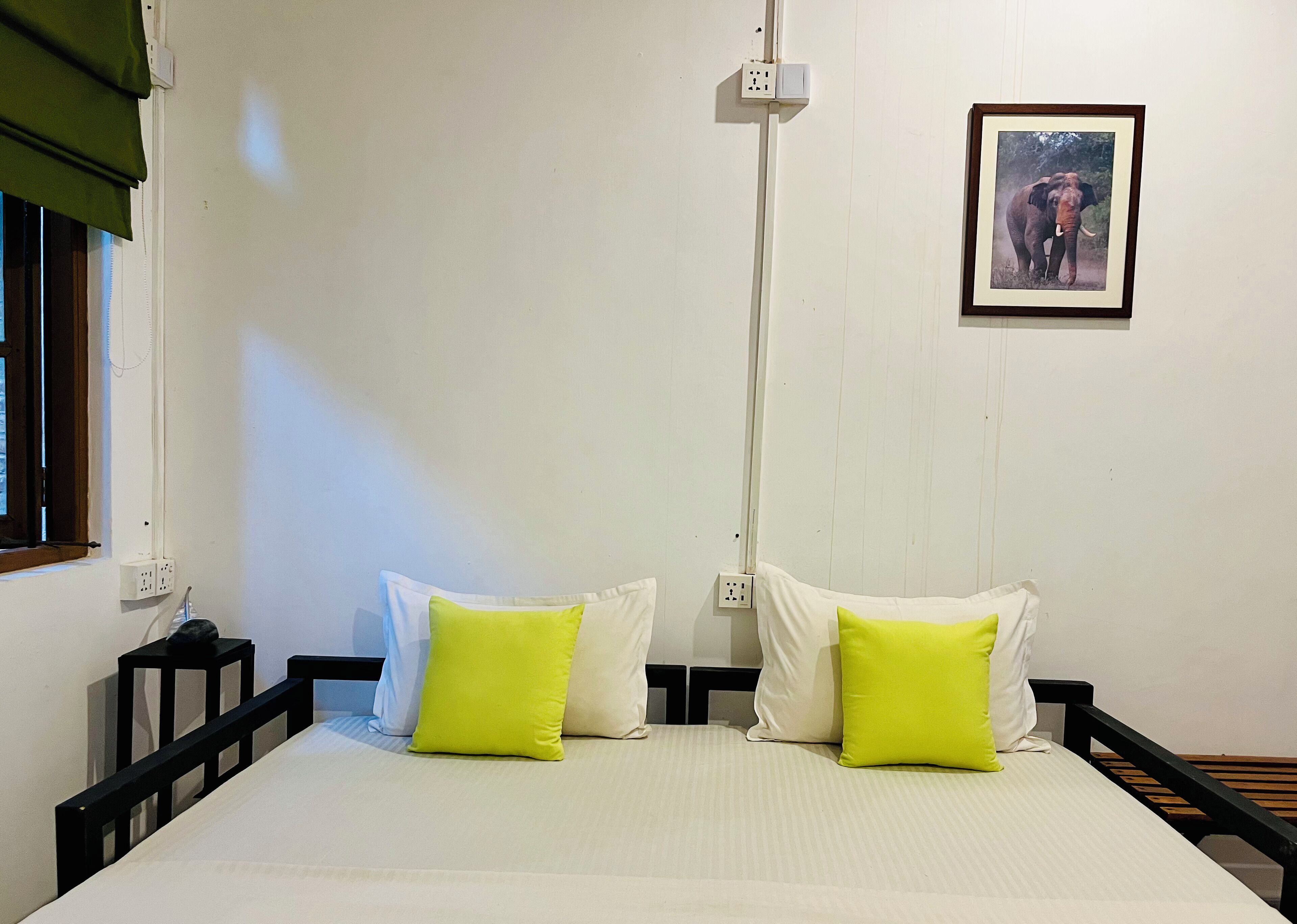 Quarto com cama, duas almofadas, um quadro na parede e janela com persianas.