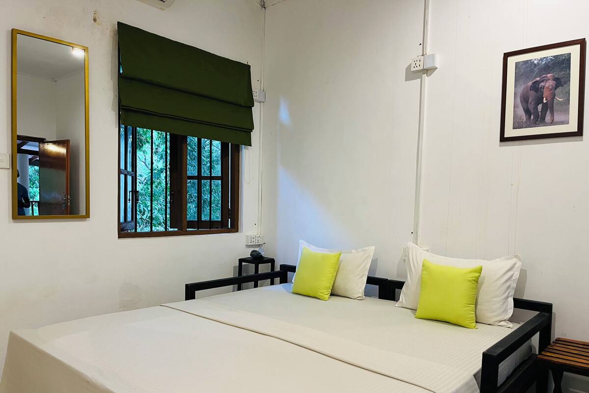 Quarto simples e limpo com cama, janela com persiana verde, espelho e quadro na parede.