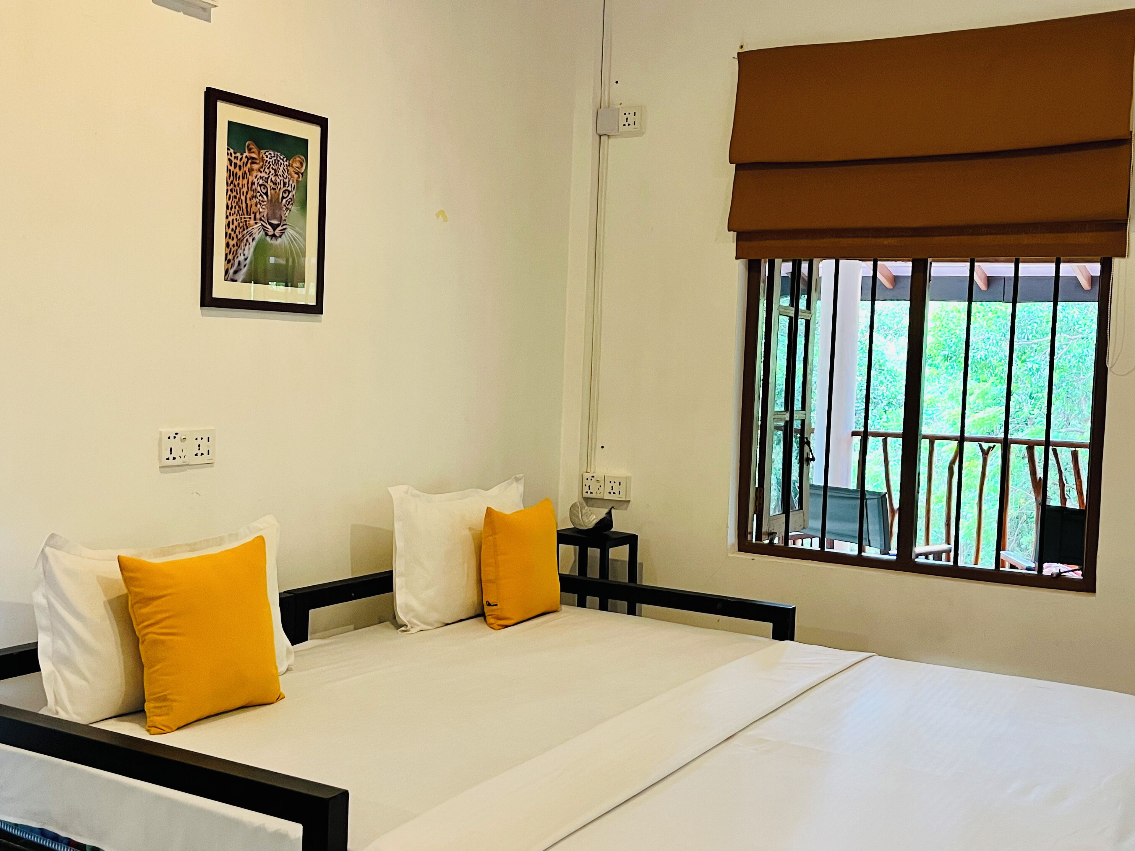 A hotel room with a bed, pillows, a nightstand, a painting, and a window with a view of greenery.