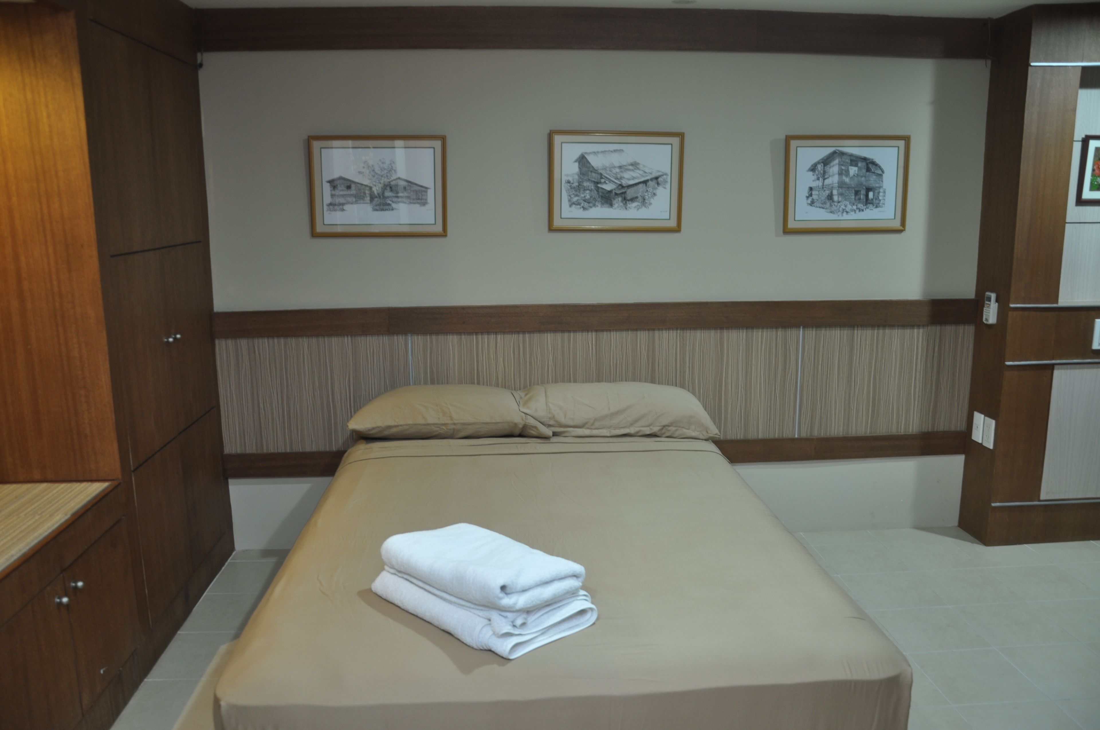 A neatly made bed with a folded white towel, a wooden wardrobe, and three framed pictures on the wall.