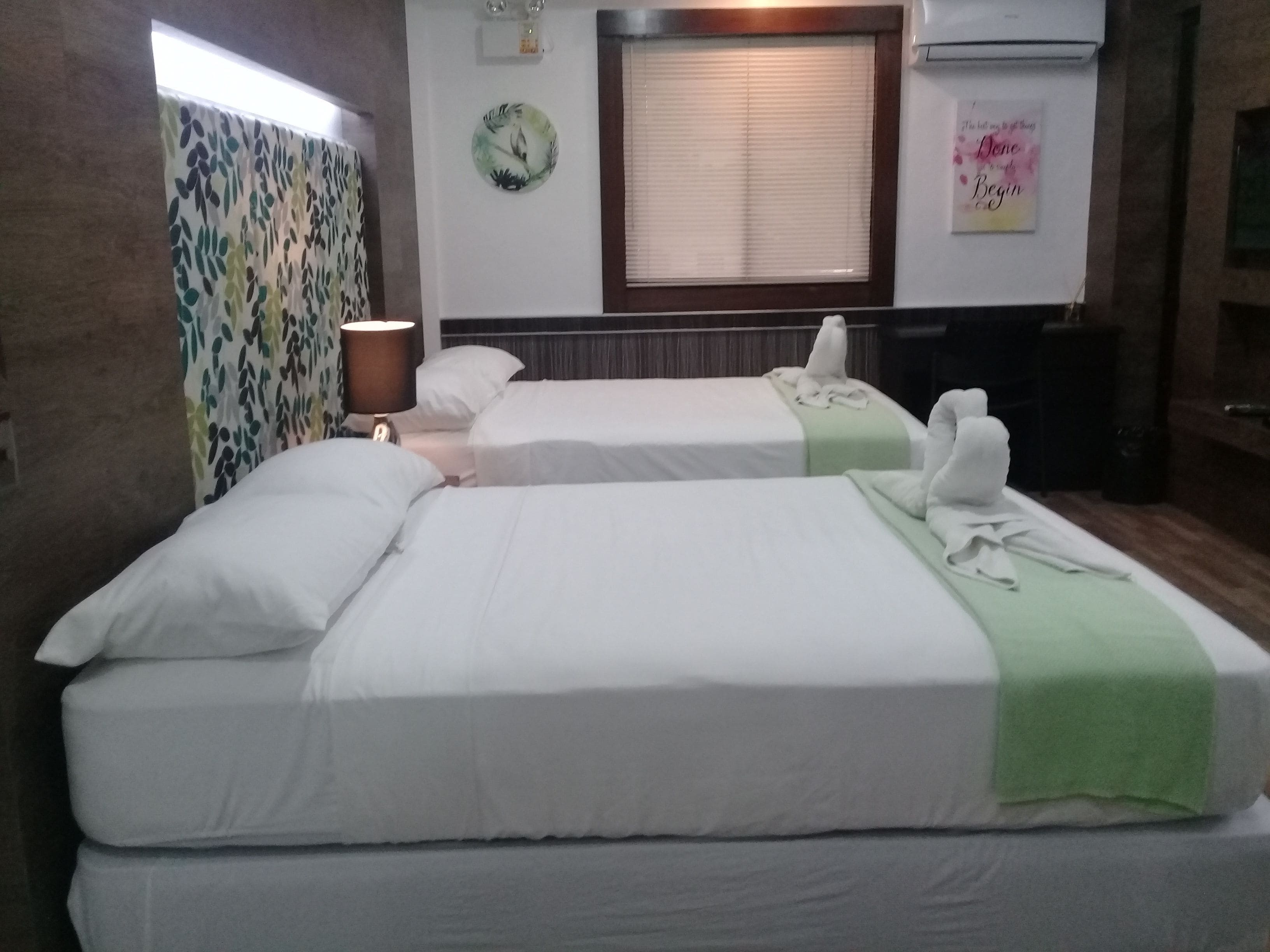 A hotel room with two beds, a window with blinds, a wall clock, and a decorative wall piece.