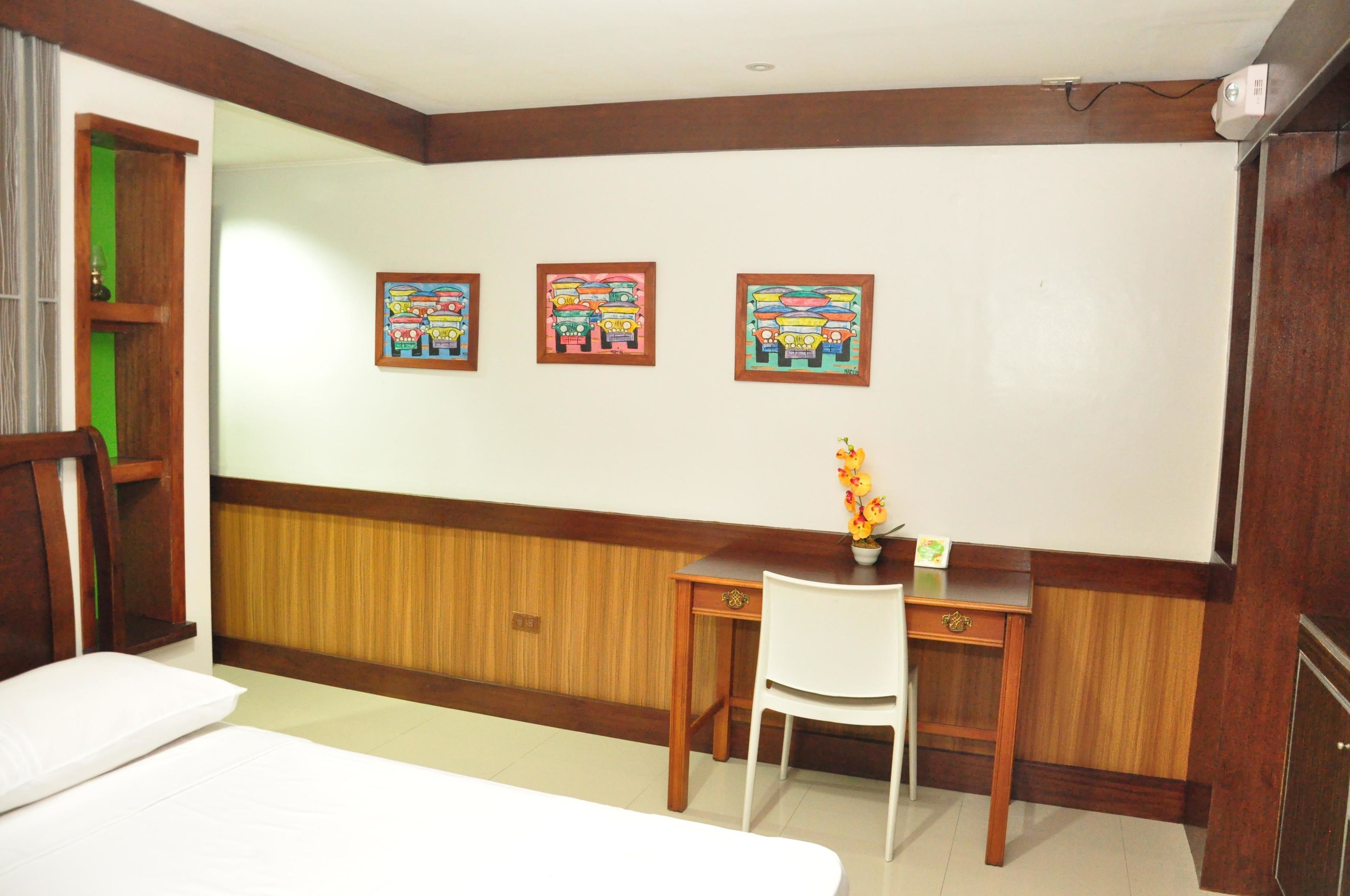 A hotel room with a bed, a desk with a chair, a small table with a vase of flowers, and three framed pictures on the wall.