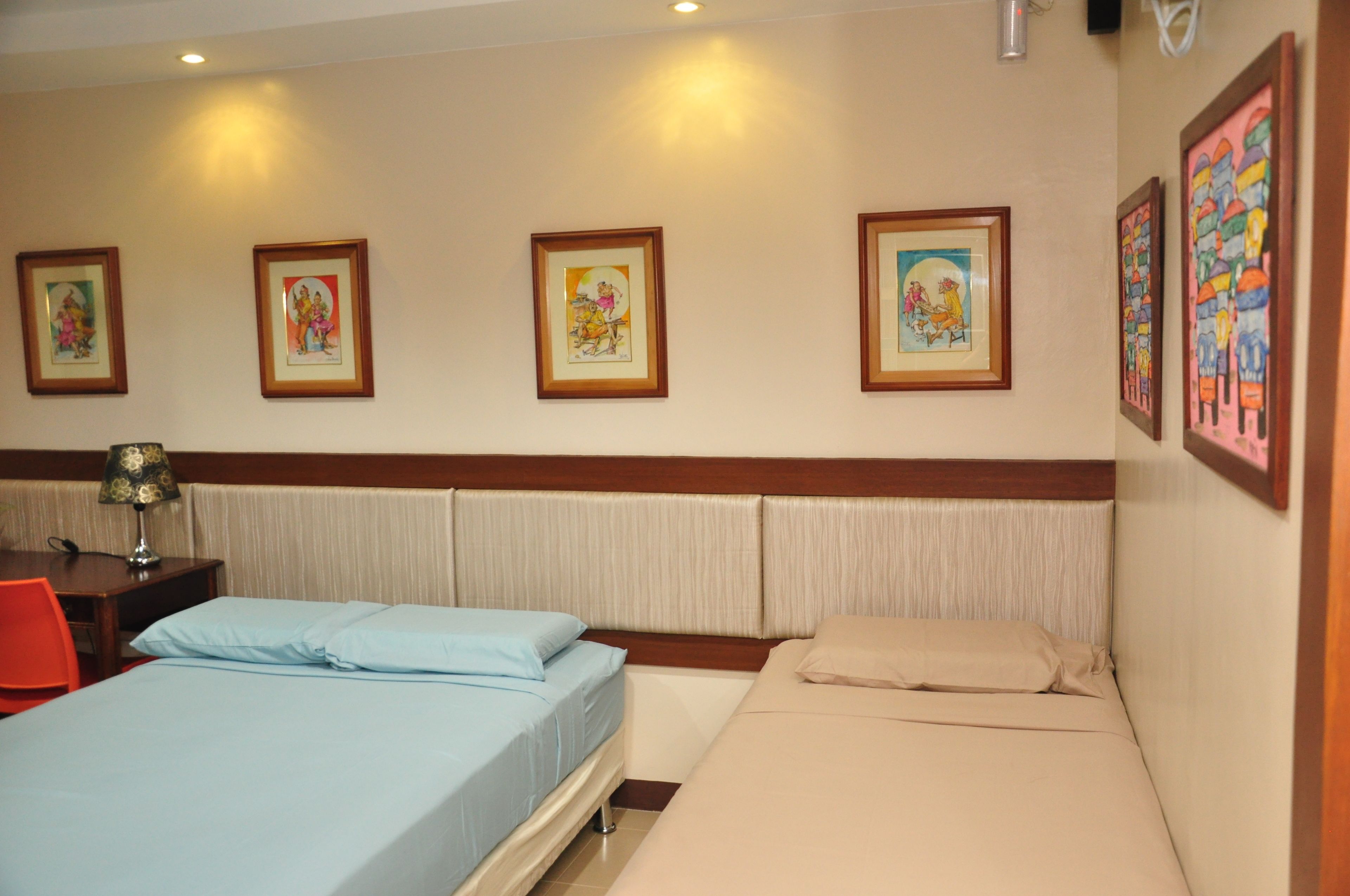 A hotel room with two beds, a desk with a lamp, and four framed pictures on the wall.